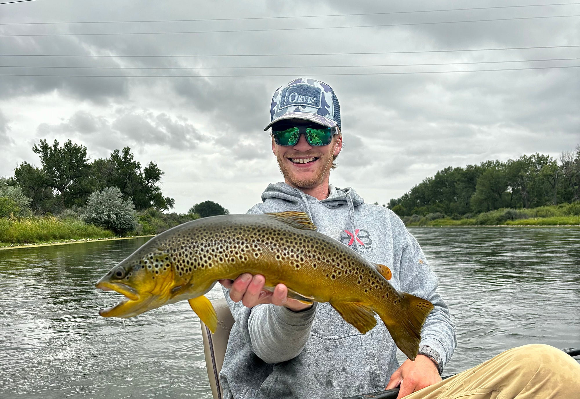 Smiling angler wearing a cap and sunglasses holding a large brown trout on a river with trees in the background