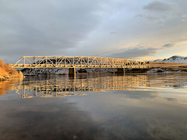 Metal truss bridge spanning calm river with reflections and distant hills under cloudy sky at sunset